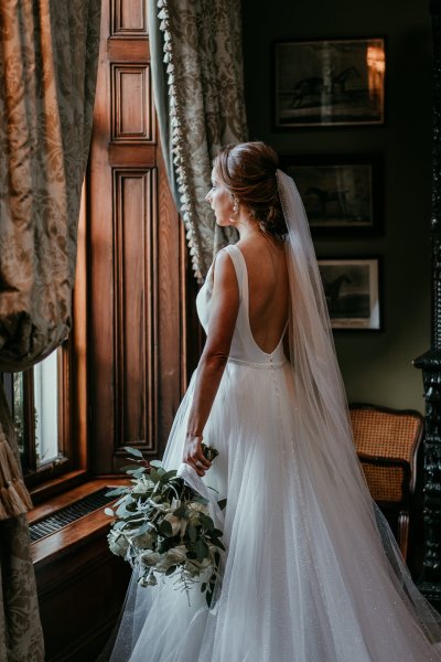 Bride stands at window looking out with bouquet in hand