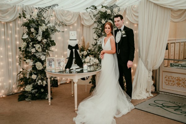 Bride and groom pose beside white wedding cake with black ribbon bow around it