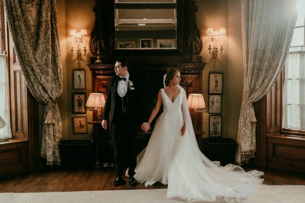 Bride and groom hold hands in manor house setting looking away