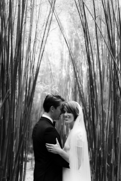 Black and white image of bride and groom standing under forest trees