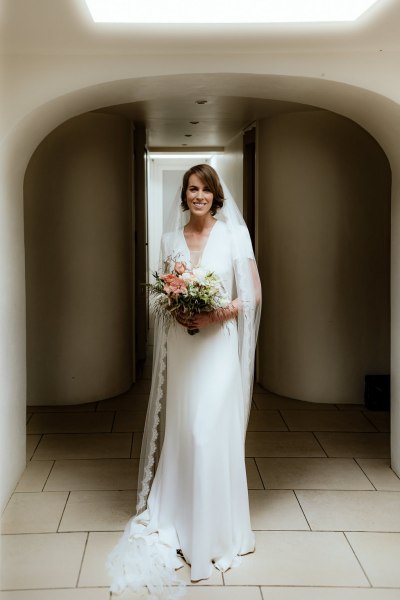 Bride stands in doorway doorframe holding bouquet smiling