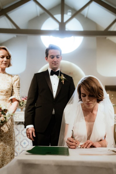 Bride signs marriage cert with groom behind