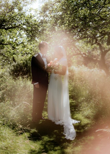 Bride and groom touch foreheads in forest setting holding bouquet of flowers sunlight