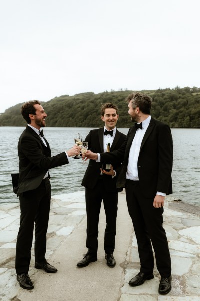 Groom and groomsmen stand on boardwalk to lake with champagne/prosecco in hand