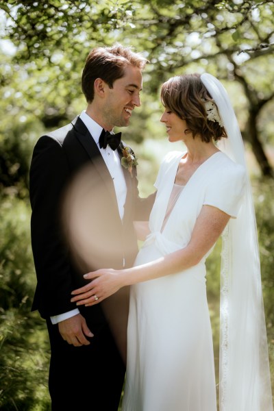 Bride and groom look at each other in forest setting holding hands sunlight reflection