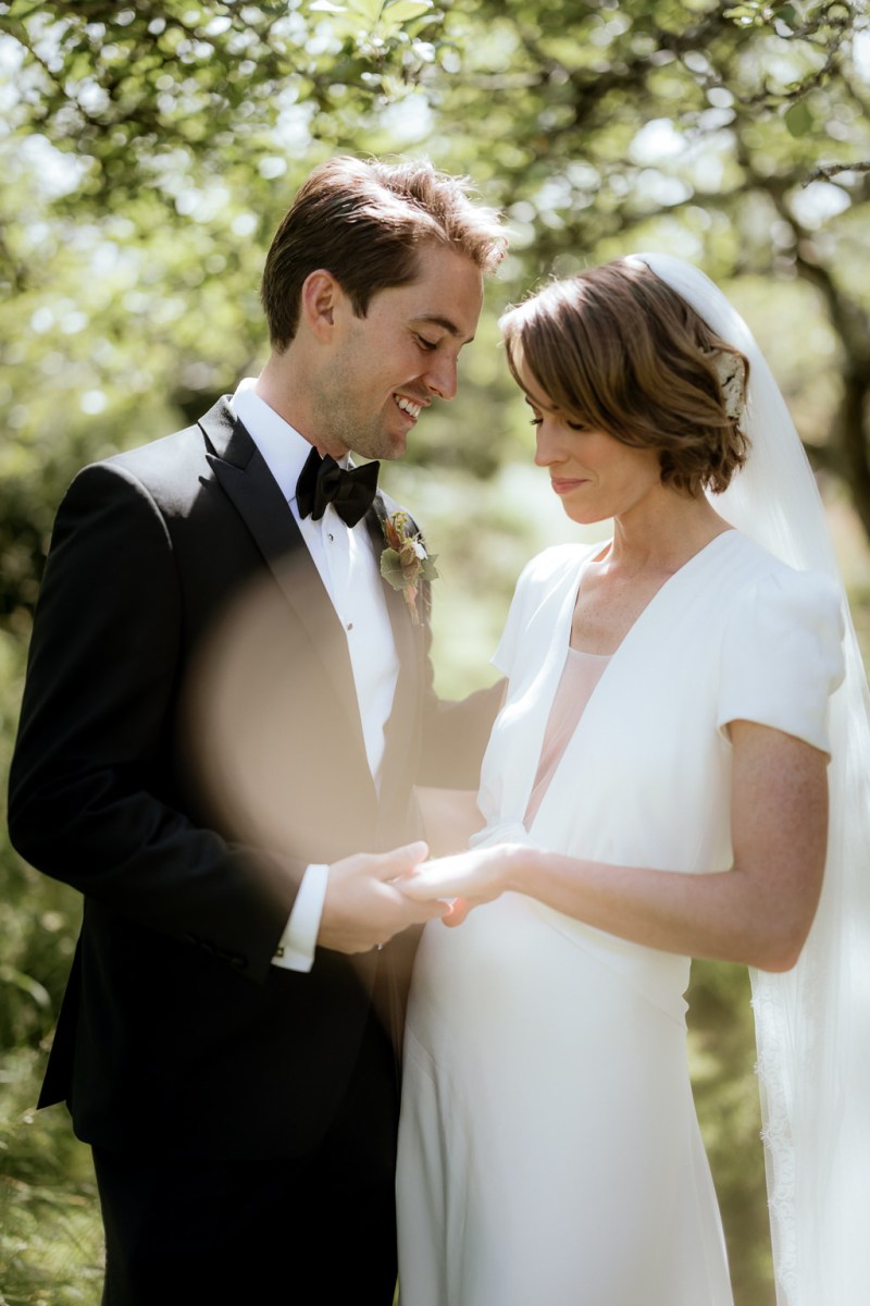 Bride and groom look at each other in forest setting holding hands sunlight reflection