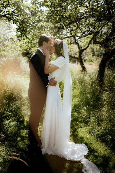 Bride and groom kiss hug embrace on grass sun shining through trees forest setting