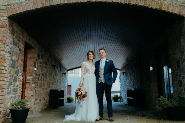 Bride and groom around the waist hug pose for the camera under bridge