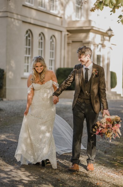 Bride and groom walk hand in hand outside wedding venue bouquet in shot