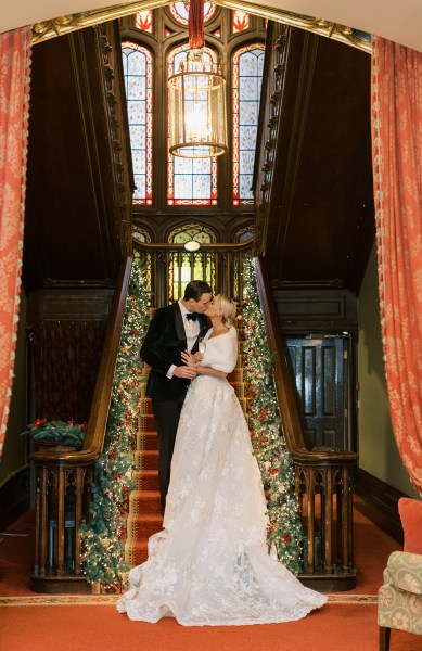 Bride and groom standing at bottom of staircase lights holly surround them kissing kiss