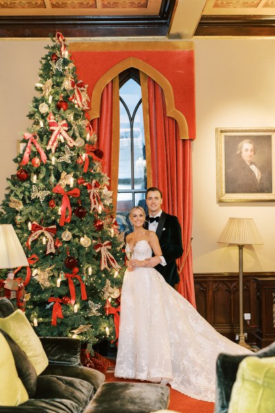 Bride and groom stand holding champagne beside the Christmas tree window in background