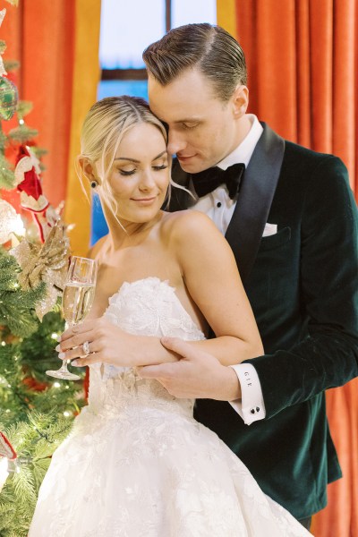 Bride and groom stand holding champagne beside the Christmas tree window in background