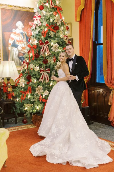 Bride and groom stand holding champagne beside the Christmas tree window in background