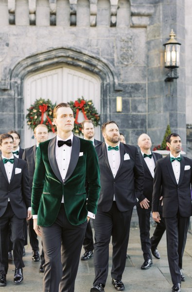 Groom stands outside exterior shot wreath hanging behind him on door with groomsmen walking behind him