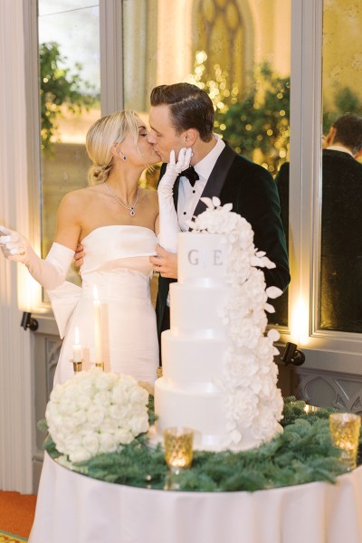 Bride from behind bow detail on dress gown and groom cutting the white wedding cake holding knife kissing kiss each other