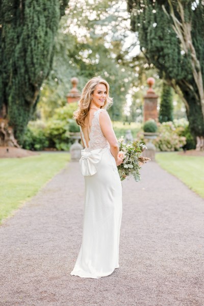 Bride shows of bow detail of bridal gown from behind standing on pathway