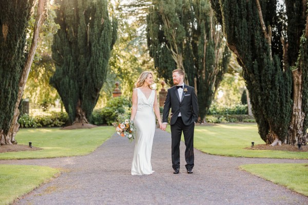 Bride and groom look at each other on pathway smiling holding hands