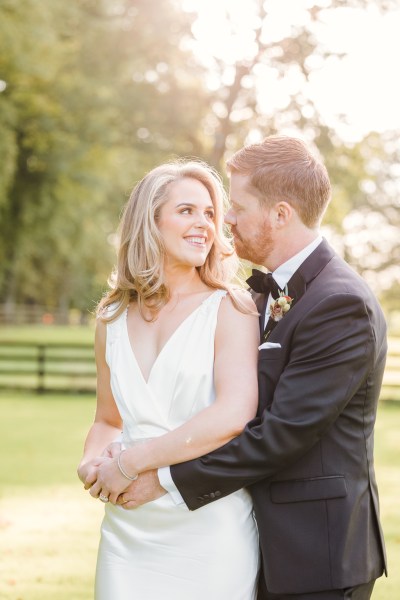 Bride and groom hold each other from behind on farm grass setting