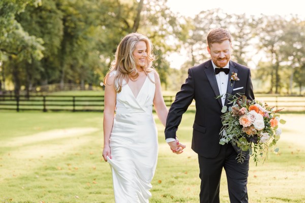 Bride and groom walk hand in hand on the pathway to wedding venue holding bouquet on grass