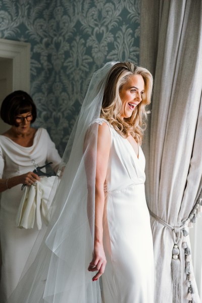 Bride wearing veil smiles in front of window