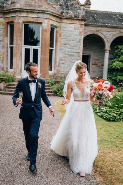 Bride and groom hold glasses of prosecco/champagne as they walk outside wedding venue