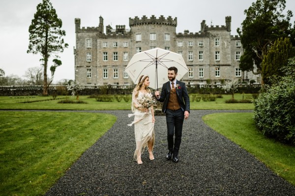 Bride and groom walk with white umbrella along pathway outside wedding venue