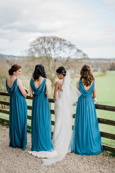 Bride and bridesmaids in blue standing beside farm fence trees and grass