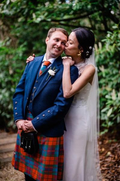 Bride hugs groom from behind and kisses his cheek in forest setting