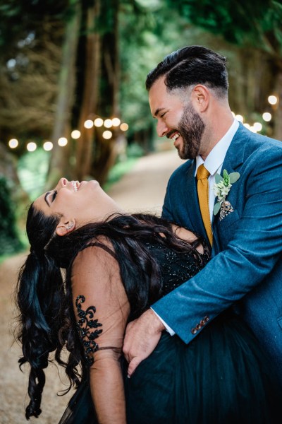 Bride and groom dance smile and laugh in park garden pathway lights in background