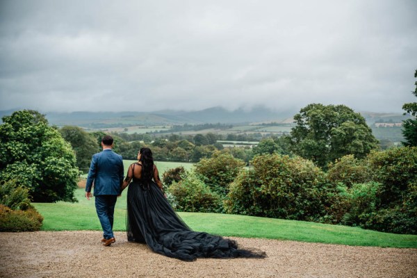 Bride and groom walk along pathway in garden long black dress train