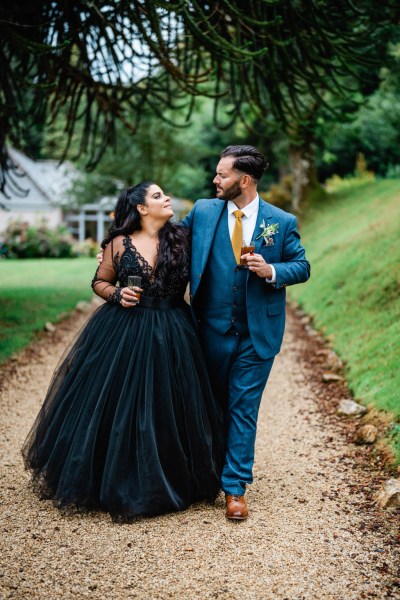 Bride and groom walk along pathway holding beverages