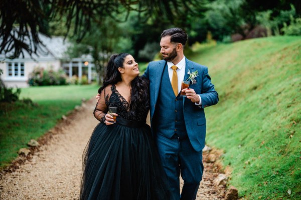 Bride and groom walk along pathway holding beverages