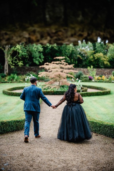 Bride and groom walking along pathway hand in hand from behind