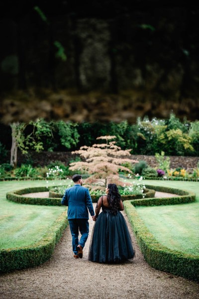 Bride and groom walking along pathway hand in hand from behind