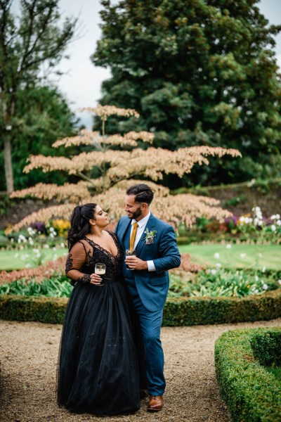 Bride and groom hold beer/Guinness beverages in garden looking at each other