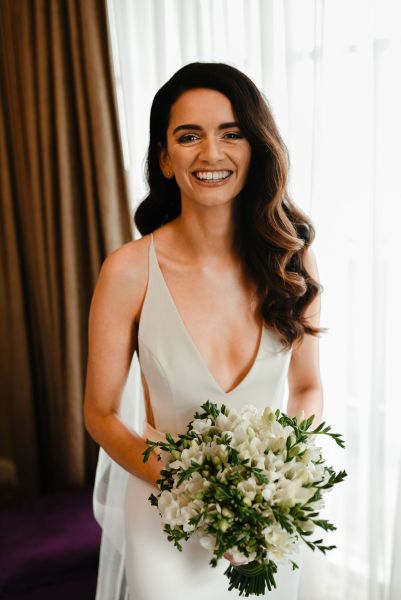 Bride stands at window smiling holding bouquet