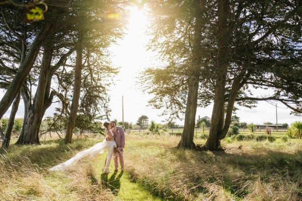 Bride and groom in forest park train dress blowing in the wind on grass wideshot sun shining trees surround couple