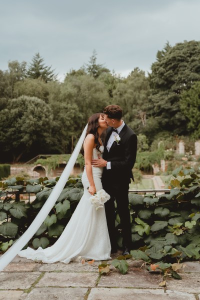 Groom places hand on brides waist and kisses in garden setting