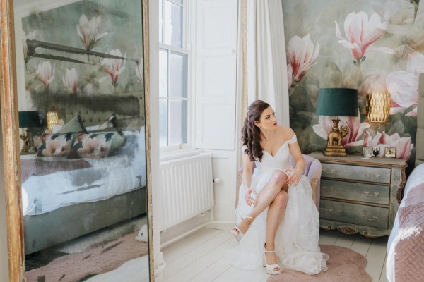 Bride sitting at window putting bridal heels/shoes on