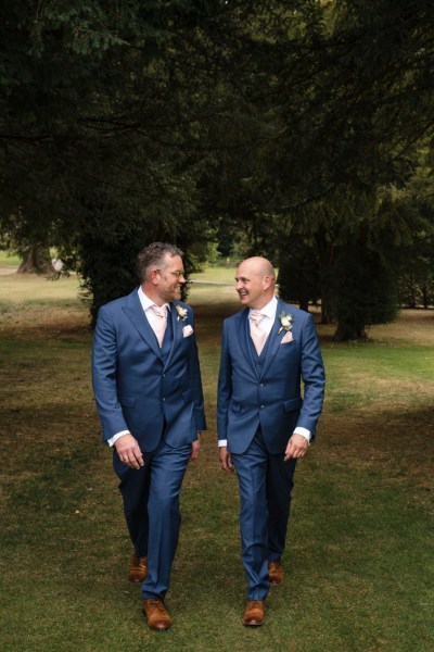 Groom and groomsman walking along grass in forest