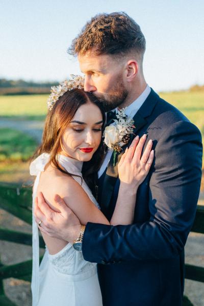 Bride and groom standing in front of fence on farm sunset facing each other hugging