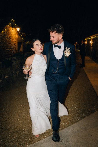 Bride and groom walk with glass of wine night time shot walking along pathway