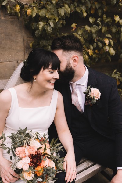 Bride and groom sitting on bench both smiling whisper in the ear