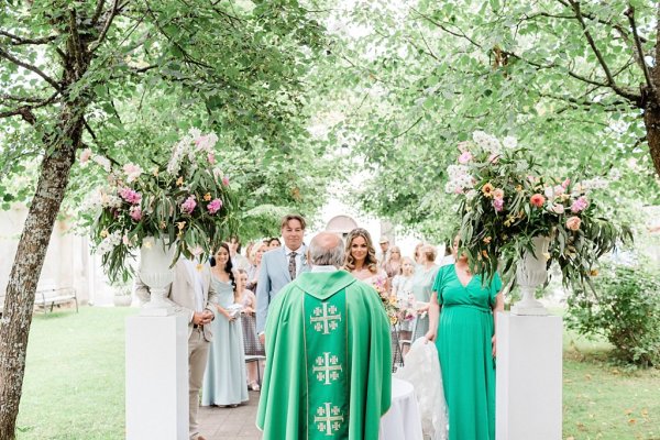 Bride and groom hold hands at alter in front of guests during ceremony priest from behind