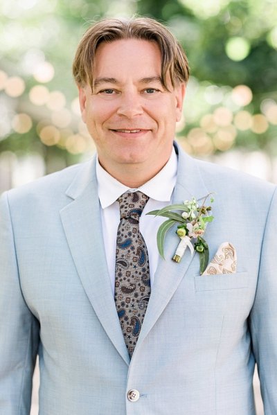 Groom on his own blue suit he poses for the camera as he stands in park forest flower detail brooch