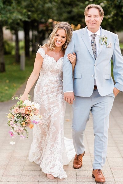 Bride and groom hold hands in forest park setting bouquet in hand flowers in background arm in arm