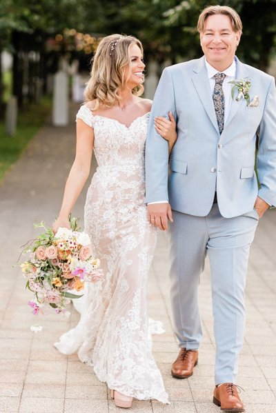 Bride and groom hold hands in forest park setting bouquet in hand flowers in background arm in arm