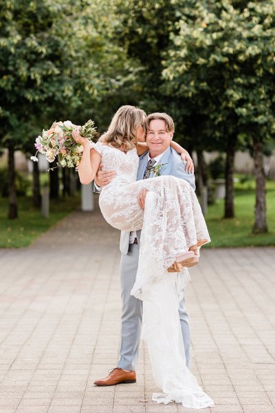 Groom picks up bride standing in park trees in background