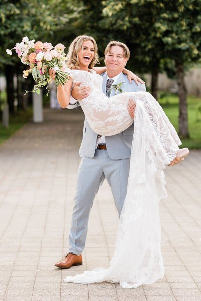 Groom picks up bride standing in park trees in background