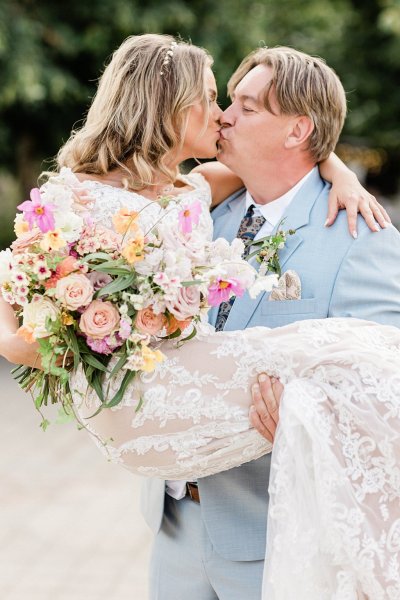 Groom picks up bride standing in park trees in background they kiss kissing